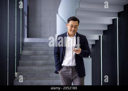 Ein fröhlicher asiatischer Geschäftsmann in Anzug und Brille, der ein Smartphone benutzt, während er eine stilvolle Treppe in einem modernen Büroambiente hinabsteigt. Stockfoto