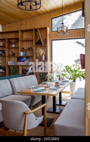 Ein rustikales Restaurant mit Holzvertäfelung, großen Fenstern, Blick auf die schneebedeckte Landschaft, Tischen, Stühlen, Kamin und Bar schaffen ein warmes Ambiente. Stockfoto