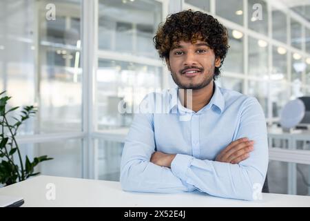 Ein junger Mann mit lockigen Haaren und einem charmanten Lächeln, der selbstbewusst in einer hellen Büroumgebung sitzt, die Arme überkreuzt und elegant in einem hellblauen Hemd gekleidet ist. Stockfoto