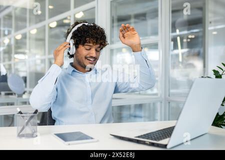 Ein fröhlicher junger Mann mit lockigen Haaren, der Kopfhörer trägt, lauscht fröhlich Musik, während er an seinem Schreibtisch tanzt, umgeben von moderner Technologie. Stockfoto