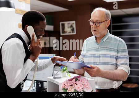 Der afroamerikanische Concierge geht ans Telefon, ein alter kaukasischer Rentner unterschreibt Reservierungsformulare an der Hotelrezeption. Während die Empfangsdame telefoniert, füllt der ältere Gast die Check-in-Formulare aus. Stockfoto
