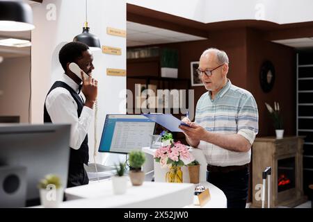 Ein alter Mann im kaukasischen Ruhestand unterschreibt Reservierungsdokumente an der Hotelrezeption, während der afroamerikanische Concierge Anrufe entgegennimmt. Elterner, männlicher Kunde, der die Check-in-Formulare ausfüllt, während der Rezeptionist telefonisch spricht. Stockfoto