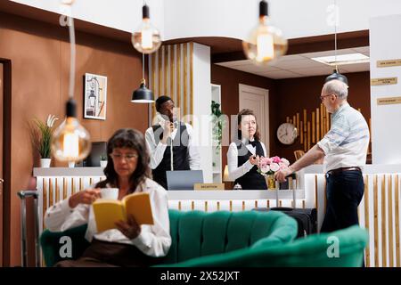 Mit Hilfe des kaukasischen Rezeptionisten in der Hotellobby, pensionierter Senior-Mann, der Koffer an der Rezeption einchecken konnte. Während ein männlicher Besucher an der Theke wartet, liest eine ältere Frau im gemütlichen Loungebereich. Stockfoto