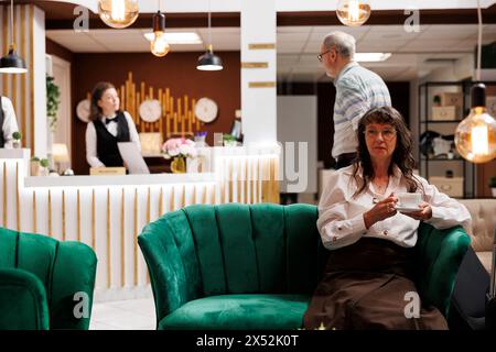 Ältere kaukasische Frau saß zum Check-in im luxuriösen Loungebereich mit einer Tasse Kaffee auf dem Sofa. Ein älterer Mann kommt in der Hotellobby an, während die alte Touristenfrau auf der Couch auf die Buchung wartet. Stockfoto