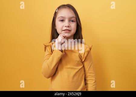Das entzückende hispanische Mädchen steht und konzentriert in reflektierendem Denken, Finger am Kinn, schaut im Zweifel auf, wundert sich über eine Idee gegen ein Gelb Stockfoto