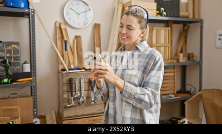 Eine lächelnde junge Frau sucht ihr Smartphone in einer gut ausgestatteten Tischlerei Stockfoto