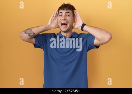 Junger hispanischer Mann, der auf gelbem Hintergrund lächelt und fröhlich einen Buh mit Händen und Gesicht zeigt, ist überrascht und begeistert Stockfoto