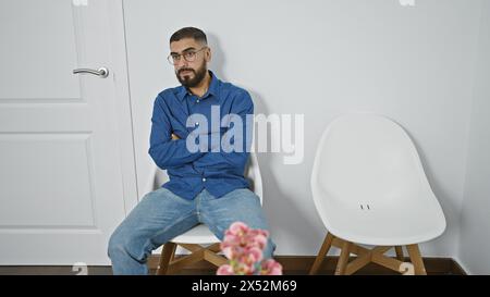 Ein bärtiger Mann sitzt überkreuzt in einem hellen, minimalistischen Zimmer mit weißem Stuhl und Inneneinrichtung, die eine ungezwungene, aber durchdachte Atmosphäre ausstrahlt. Stockfoto