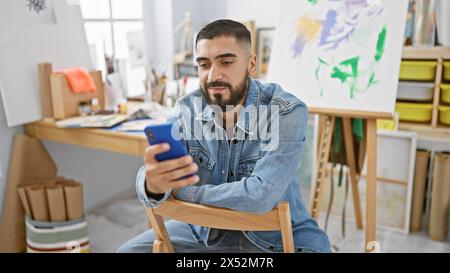 Hübscher junger Mann mit Bart in einer Jeansjacke mit Smartphone in einem Kunststudio Stockfoto