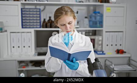 Eine nachdenkliche Frau in einem Labor, die Handschuhe trägt, während sie ein Handbuch liest, umgeben von wissenschaftlicher Ausrüstung. Stockfoto