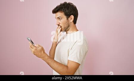 Ein überraschter hispanischer Mann mit Bart sieht sein Handy vor rosa Hintergrund an. Stockfoto