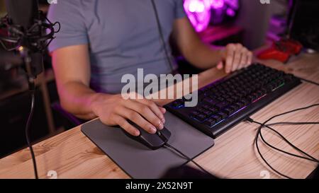 Ein junger hispanischer Mann benutzt eine Computermaus und Tastatur in einem dunklen Spielzimmer mit violetten LED-Lichtern Stockfoto