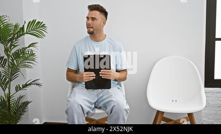 Ein junger hispanischer Mann mit Bart sitzt in einem Innenraum und hält ein Klemmbrett und sorgt so für eine professionelle, aber entspannte Atmosphäre. Stockfoto