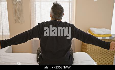 Ein reifer hispanischer Mann mit grauen Haaren und Bart dehnt sich in seinem Schlafzimmer und beginnt seine Morgenroutine. Stockfoto