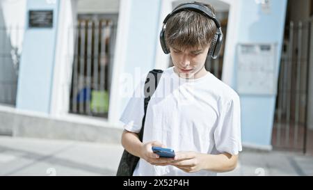 Ein junger kaukasischer Teenager in einem weißen T-Shirt benutzt ein Smartphone mit Kopfhörern auf einer städtischen Straße. Stockfoto