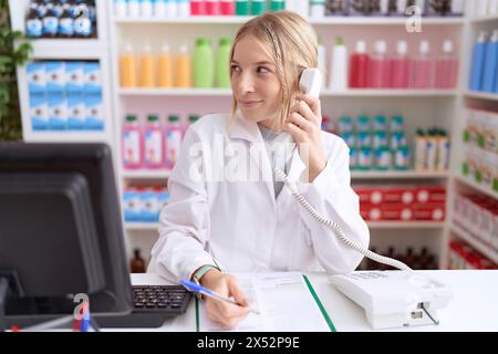 Junge kaukasische Frau, die in einer Apotheke arbeitet und am Telefon spricht, mit einem Lächeln auf dem Gesicht und einem natürlichen Ausdruck zur Seite schaut. Lachend Co Stockfoto