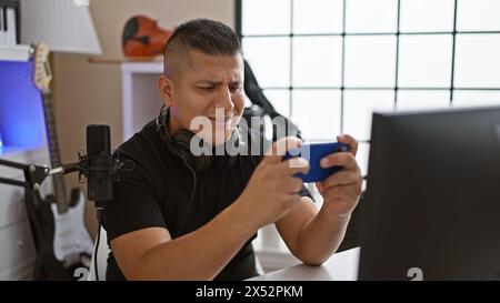 Fokussierter junger latin-man-Streamer, der digitale Spiele auf dem Smartphone in einem lebendigen Musikstudio spielt und in die Welt der Streaming-Unterhaltung eingetaucht ist Stockfoto