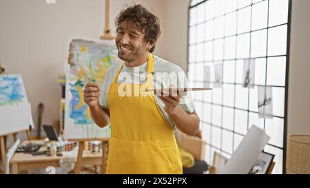 Ein fröhlicher bärtiger Mann in einer gelben Schürze malt auf Leinwand in einem hellen Kunststudio und verkörpert Kreativität und Lernen. Stockfoto