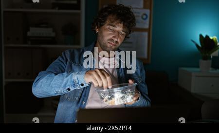 Ein junger hispanischer Mann in einer lässigen Jeansjacke isst Salat während einer späten Nacht im Büro. Stockfoto