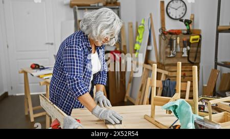 Eine reife Frau arbeitet aufmerksam in einem Tischlerstudio, umgeben von Holzmöbeln und Werkzeugen. Stockfoto