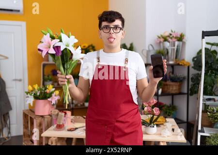 Junger, nicht binärer Mann, der im Blumenladen arbeitet, zeigt den Smartphone-Bildschirm, der die Zunge herausragt, glücklich mit lustigem Ausdruck. Stockfoto