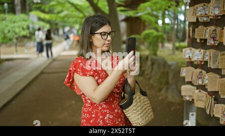 Wunderschöne hispanische Frau in Brille, die Gebetstafeln im japanischen Gotokuji-Tempel festnimmt und die Tradition durch ein Smartphone-Objektiv bewahrt Stockfoto