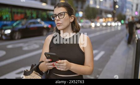 Schöne brünette hispanische Frau in Brille, vertieft in ernsthafte Smartphone-Gespräche auf madrids städtischen Straßen Stockfoto