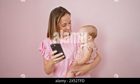 Lächelnde Mutter hält das Baby selbstbewusst in den Armen, während sie am Telefon Nachrichten sendet, und genießt ihre schöne Bindung über einem rosa isolierten Hintergrund Stockfoto