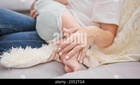 Liebevolle Mutter, die zu Hause auf dem Wohnzimmer-Sofa sitzt und die Beine ihrer liebenden Tochter berührt, um eine echte Familienbindung zu schaffen. Stockfoto
