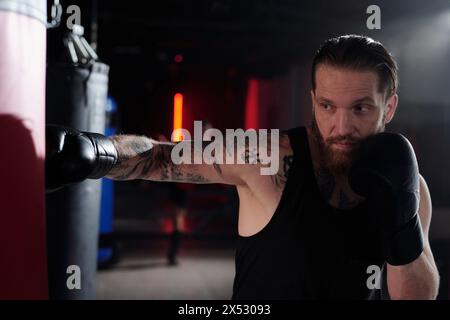 Junger Mann mit Tätowierungen am muskulösen Arm und auf der Brust, der während des Trainings im Fitnessstudio oder im Boxring einen Boxsack schlägt Stockfoto