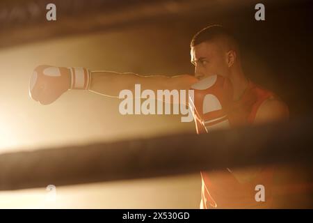 Junger Boxer, der den linken Arm im Ellenbogen am Gesicht gebeugt hält, während er den Rivalen mit der rechten Hand im Boxhandschuh während des Trainings am Ring tritt Stockfoto