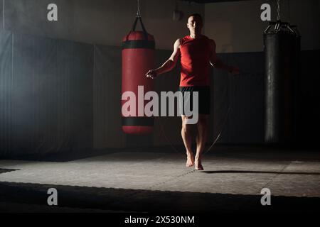 Junger Barfußsportler in roter Weste und schwarzer Shorts, der mit Seilspringen auf dem Boden des Fitnessstudios mit zwei Boxsäcken springt Stockfoto
