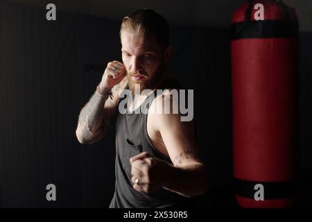 Licht fällt auf den vorderen Teil eines jungen muskulösen Mannes mit Tätowierungen auf Armen und Brust, die vor dem Boxsack stehen und einen Kick üben Stockfoto