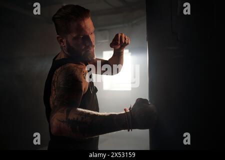 Junger muskulöser Mann mit Tätowierungen an Armen, Schultern und Brust, der Schlafsack gegen Sonnenlicht schlägt, das durch das Fenster des Fitnessstudios eindringt Stockfoto