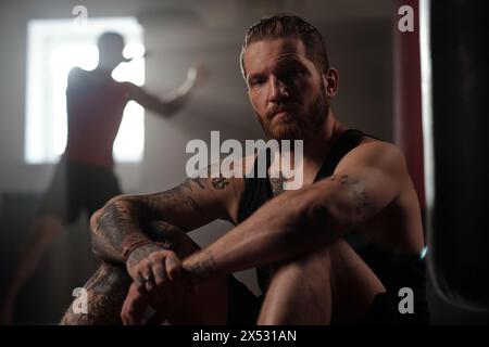 Brutaler Sportler mit Tätowierungen auf Beinen, Armen, Schultern und Brust, der auf dem Boden des Fitnessstudios sitzt und gegen den Kerl, der den Boxsack tritt Stockfoto