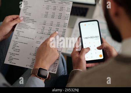 Hoher Handwinkel einer jungen, nicht erkennbaren Frau, die auf die Summe in der Finanzrechnung zeigt, während ihr Mann Berechnungen auf dem Smartphone macht Stockfoto