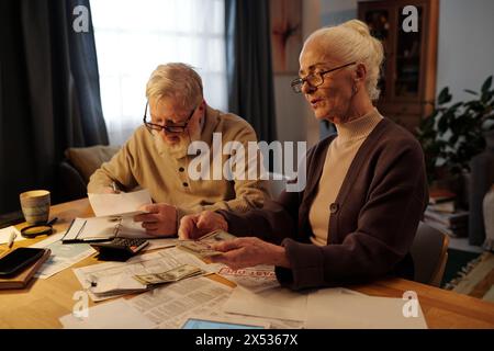 Frau mit weißem Haar, die am Tisch sitzt und Dollarscheine über bezahlte und unbezahlte Finanzrechnungen gegen ihren Mann zählt, der Notizen macht Stockfoto