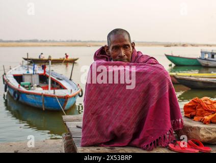 Mann, der mit Armen auf hochgezogenen Knien sitzt, umwickelt von einer kastanienbraunen Decke neben dem Ganges bei Varanasi, Indien. Stockfoto