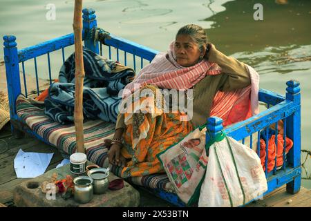 Hindu-Frau sitzt auf einer blau bemalten Holzbank mit Puja-Gegenständen vor ihr neben dem Ganges in Varanasi, Indien. Stockfoto