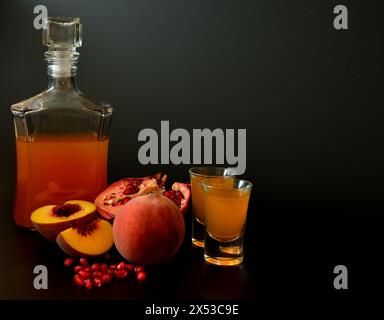 Granatapfel-Pfirsichlikör, starker hausgemachter Alkohol in einer Glasflasche und zwei Gläser auf schwarzem Hintergrund, neben reifem Obst. Nahaufnahme. Stockfoto