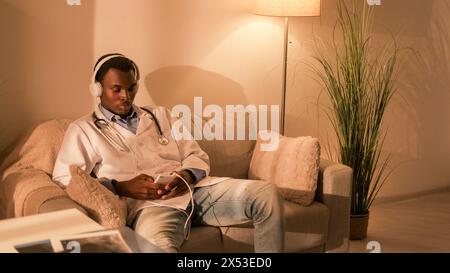 Arztbesuch. Spielzeuge für Freizeit. Internet-Erholung. Müde entspannter männlicher Arzt, der Musik in den Kopfhörern auf dem Telefon auf der Couch im modernen Abendinnenraum mit freiem Platz hört. Stockfoto