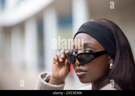 Ein Nahporträt einer stylischen Afroamerikanerin, die ihre übergroße Sonnenbrille im Freien anpasst. Ihre Kleidung ist lässig und doch schick, ergänzt durch dezentes Make-up und ein schwarzes Stirnband. Der verschwommene urbane Hintergrund deutet auf eine städtische Umgebung hin und verleiht dem Bild eine kosmopolitische Atmosphäre. Stilvolle Afroamerikanische Frau, Die Sonnenbrille Im Freien Verstellt. Hochwertige Fotos Stockfoto