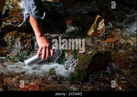 Wasser fließt über Felsen. Ein Tramper oder Wanderer lehnt sich nach unten und füllt eine Wasserflasche aus Edelstahl aus dieser Süßwasserquelle Stockfoto