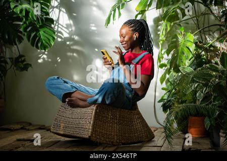 Fröhliche schwarze Frau, die sich im Innengarten entspannt. Afroamerikaner mit Smartphone im sonnigen Garten Stockfoto