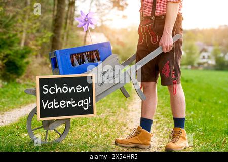 7. Mai 2024: Mann in Bayerischer Lederhose schiebt einen Bollerwagen am Vatertag mit einem Schild und dem Gruß: Hab eine schöne Vatertag FOTOMONTAGE *** Mann in bayerischer Lederhose schiebt einen Bollerwagen am Vatertag mit einem Schild und dem Gruß: Schön Vatertag FOTOMONTAGE Stockfoto