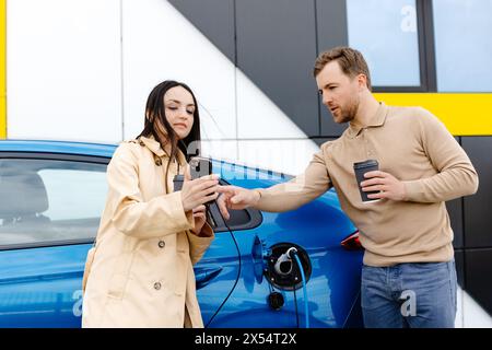 Ein junges Paar, das mit einem Elektroauto unterwegs ist und an der Ladestation hält. Freund, der das Kabel zum Aufladen einsteckt. Ein Mann, der mit einer Freundin redet Stockfoto