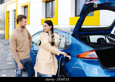 Ein junges Paar, das mit einem Elektroauto unterwegs ist und an der Ladestation hält. Eine Freundin, die das Ladegerät hält, das Auto ansieht und lächelt. Stockfoto