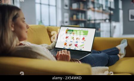 Schöne kaukasische Frau verwendet Laptop-Computer für die Bestellung der Lieferung von frischem Obst und Gemüse. Sie ist im Wohnzimmer zu Hause. Freiberuflicher Spezialist, der von zu Hause aus arbeitet, im Internet surft. Stockfoto