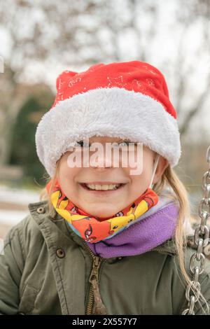 Freundliches, hübsches Mädchen auf einem Spielplatz mit einem schicken roten Weihnachtsmann-Hut. Im Winter, kurz vor Weihnachten, gibt es große Begeisterung über die Geschenke. Stockfoto