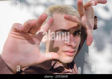 Ein junger lateinmann mit einem rasierten Gesicht posiert für ein Bild. Er trägt ein braunes Hemd und hat seine Hände vor dem Gesicht. Das Bild hat einen Casu Stockfoto